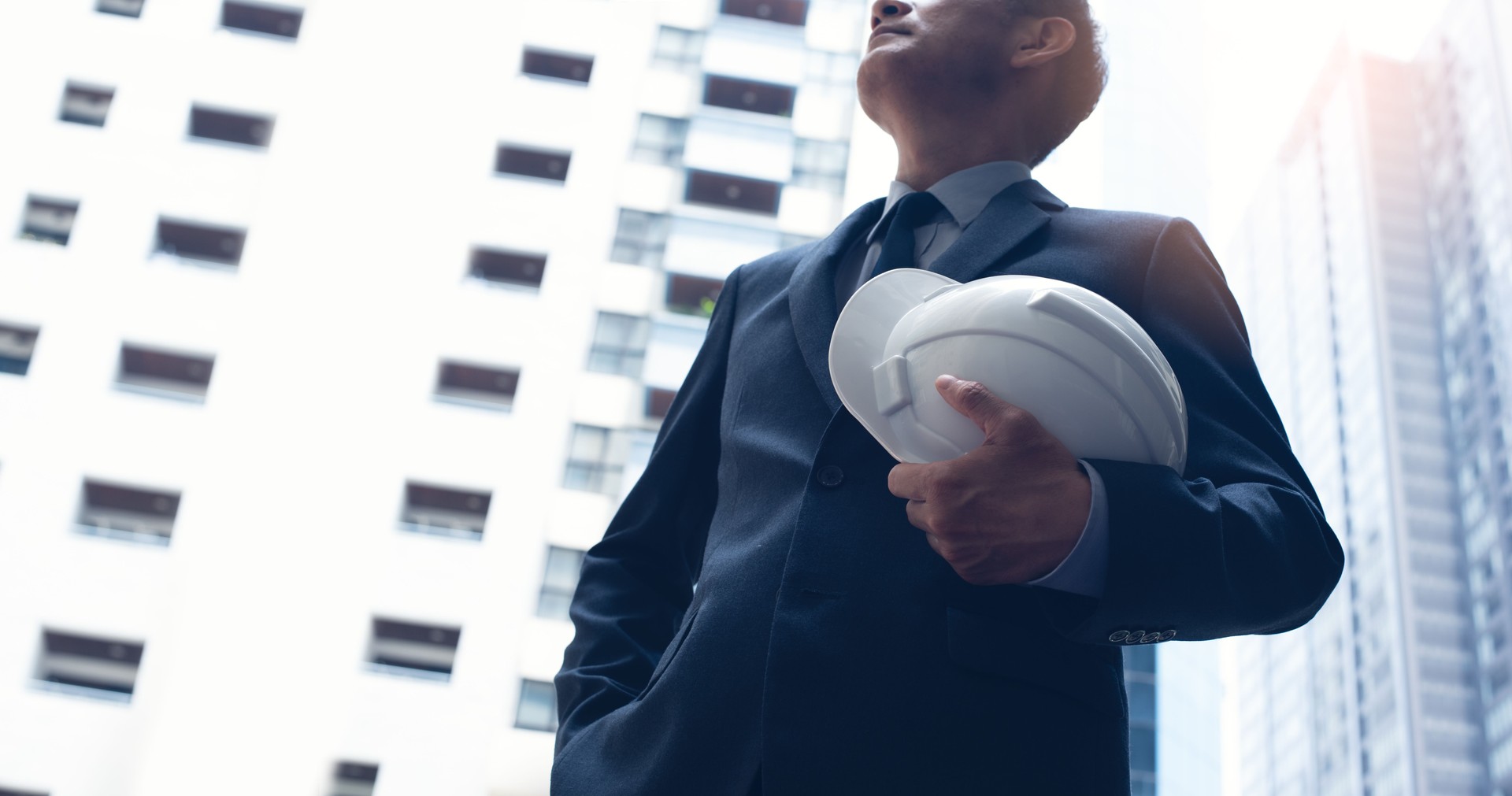 Engineer with construction helmet standing in front of skyscraper buildings in the city with sunlight, close up